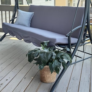 May include: A gray outdoor swing with a matching cushion and a light blue pillow. The swing is on a wooden deck, and a potted plant in a woven basket sits in front of it. The swing has a metal frame.
