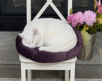 May include: A white cat curled up asleep on a round, deep purple cushion atop a white chair. A bouquet of pink flowers in a silver bucket sits to the right. The chair has an X-shaped back.