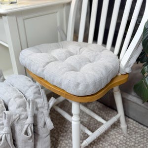 May include: A light gray tufted chair cushion with a textured fabric finish and fabric ties, secured to a wooden chair seat. The cushion features a square shape with rounded corners and multiple stitched indentations across the surface.
