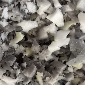May include: Close-up of a pile of shredded foam in various shapes and sizes. The foam pieces are primarily white, gray, and a pale yellow-green. The texture appears soft and porous, suggesting use in cushioning or padding.