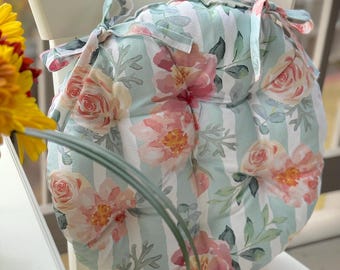 May include: Round chair cushion with a floral pattern. The cushion features a light blue and white striped background with watercolor-style pink and peach roses and green leaves. The cushion has ties for securing it to a chair.