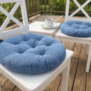 May include: Two round, tufted seat cushions in a textured blue fabric, each placed on a white wooden chair. The cushions feature a button-tufted design and a thick, padded construction, providing a soft surface for seating on the chairs.