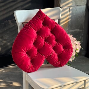 May include: A heart-shaped, red velvet cushion with a tufted design, resting on a white wooden chair. The cushion's plush texture and vibrant color create a striking visual contrast. The chair is positioned in front of a neutral background.