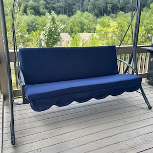 May include: A dark blue outdoor swing with a metal frame. The swing has a padded seat and backrest, and a scalloped edge on the seat cover. The swing is on a wooden deck, with trees in the background.