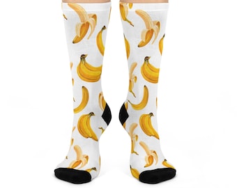 May include: A pair of white crew socks featuring an all-over repeating pattern of yellow bananas in various stages of peeling. The socks have solid black toe and heel sections, providing a distinct contrast to the white fabric and yellow fruit print.