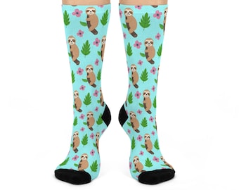 May include: A pair of light blue crew socks featuring a repeating pattern of brown sloths hanging from green leaves and small pink flowers. The socks have solid black heels and toes, providing a distinct contrast to the colorful patterned fabric.
