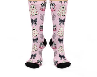 May include: A pair of light pink crew socks featuring a repeating pattern of white ghosts with pink flower accents, pink pumpkins topped with black and white checkered bows, black lace-style bows, and black spiderwebs, finished with black toe caps.