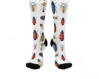 May include: A pair of white crew socks featuring an all-over print of various colorful beetles and insects in shades of red, blue, yellow, and brown. The socks have solid black heels and toes, providing a distinct contrast to the white fabric.