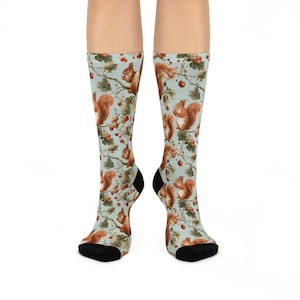 May include: A pair of crew-length socks featuring a light blue background with an all-over pattern of brown squirrels perched on tree branches among green leaves and small red berries. The socks have solid black toe and heel sections.