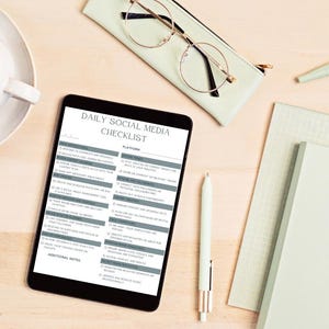 Może przedstawiać: Czarny tablet wyświetlający "Daily Social Media Checklist" na jasnej drewnianej powierzchni. Lista kontrolna zawiera sekcje dotyczące działań i platform. Inne elementy to filiżanka kawy, okulary, długopis i notatnik.