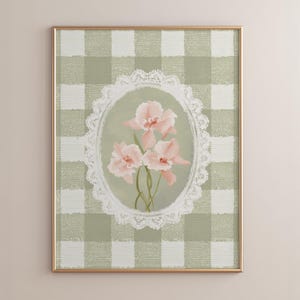 Pode incluir: Impressão floral emoldurada com orquídeas rosa dentro de um oval de renda branca contra um fundo xadrez verde e branco. A obra de arte está em uma moldura dourada, criando uma estética vintage. Medidas em centímetros.