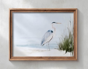 May include: A framed wall art print featuring a painting of a great blue heron standing on a sandy beach near tall green grass. The bird has grey and white feathers with a long neck and a pointed beak. The background shows a soft, misty shoreline.