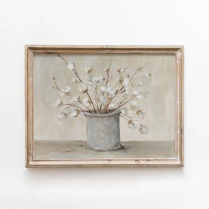 May include: A framed painting depicting a bouquet of white cotton stems arranged in a cylindrical gray vase. The artwork features a neutral color palette with soft beige and cream tones in the background, enclosed in a light-colored wooden frame.