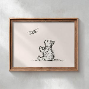 May include: A framed wall art print featuring a black ink sketch of a seated bear looking up at a small aeroplane flying above. The artwork is presented in a simple wooden frame with a light beige background, capturing a classic illustrative style.