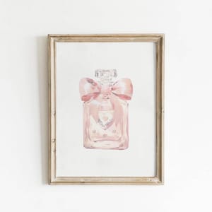 Könnte beinhalten: Aquarellmalerei einer rosa Parfümflasche mit einer großen Schleife und einem herzförmigen Design. Das Kunstwerk ist in einem goldfarbenen, rechteckigen Rahmen vor einem weißen Hintergrund gerahmt.