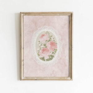 May include: A framed artwork featuring a bouquet of pink roses within an oval lace design. The roses are surrounded by green leaves and small white flowers, set against a textured pink background. The frame is a light gold colour.