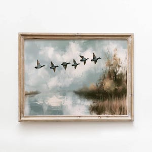 May include: A framed landscape painting depicting a flock of five ducks flying across a cloudy sky above a calm river. The scene features muted tones of blue, grey, and earthy brown, with tall grasses along the riverbank on the right side.