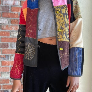 May include: A colourful patchwork jacket with embroidered details and yellow buttons. The jacket is made of different fabrics and textures, including velvet, corduroy, and cotton.