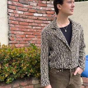 Può includere: Una persona indossa un blazer marrone e nero con un motivo a fantasia, una camicia nera e jeans marroni. Il blazer ha un tessuto strutturato e una chiusura con bottone.