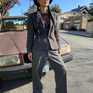 May include: A woman wearing a gray herringbone blazer and matching pants, a black hat, and sunglasses. She is standing in front of a car.