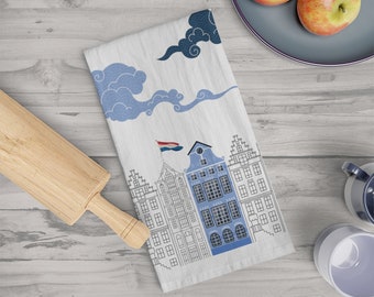 May include: White kitchen towel featuring a blue and white illustration of Dutch houses with a waving flag. The towel also has blue cloud designs. A wooden rolling pin and two apples are in the background.