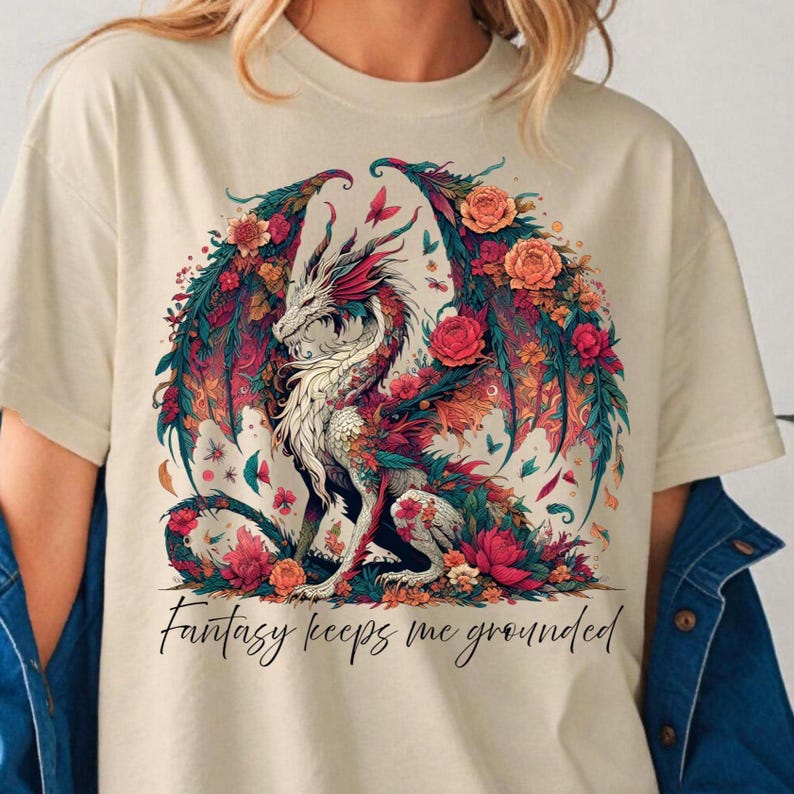 May include: Cream-colored t-shirt featuring a detailed illustration of a dragon with floral wings and accents. The dragon is white, red, and teal, surrounded by flowers. The text "Fantasy keeps me grounded" is written in cursive below the dragon.