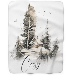 Puede incluir: Una manta blanca con una ilustración en acuarela de una cabaña cubierta de nieve entre árboles de hoja perenne. La palabra "Cozy" está escrita en cursiva en la parte inferior. Hay pájaros volando en el cielo.