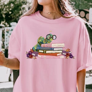 Peut inclure: Un t-shirt rose clair avec une illustration représentant une chenille verte portant des lunettes rondes, posée sur une pile de livres colorés. Le livre central est bleu avec le mot WISDOM inscrit sur la tranche. Des fleurs entourent les livres.