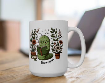Può includere: Tazza in ceramica bianca con un verme libro verde che indossa gli occhiali e legge un libro. Il verme libro è circondato da fiori e piante. La tazza dice "Bookworm" in basso.