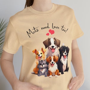 May include: A light tan t-shirt featuring a graphic of five smiling dogs and a cat. The text "Mutts need love too!" is written above the animals with two red hearts. The shirt is made of a soft material.