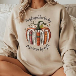 Puede incluir: Sudadera beige con un diseño de calabaza hecho de libros. La calabaza tiene un tallo verde y hojas naranjas. El texto "Pumpkin patches by day, page-turner by night" está escrito en una fuente de escritura.