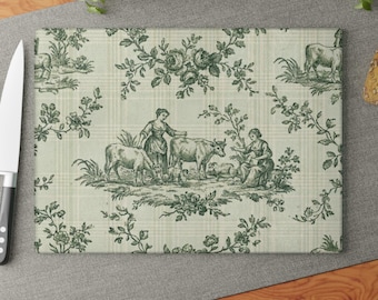 May include: Rectangular cutting board with a green and white toile pattern. The design features pastoral scenes with figures, cows, and floral elements. The board has a plaid background and is surrounded by a knife and wooden surface.
