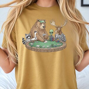 May include: Mustard-coloured t-shirt with a humorous poker scene. A bear, moose, alien, raccoon, and duck are around a green poker table. The bear wears a green visor, and the duck wears a hat. The graphic is a cartoon-style design.