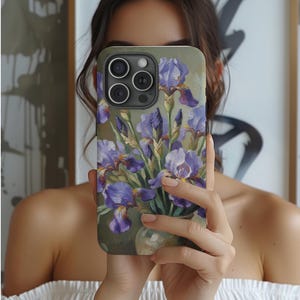 Puede incluir: Funda de teléfono con una pintura de lirios morados en un jarrón. La obra de arte tiene un estilo suave y pictórico con tonos de morado, verde y blanco. La funda es de color verde oliva y tiene tres lentes de cámara.