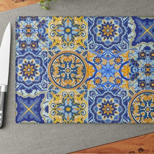 May include: A rectangular cutting board with a vibrant, patterned design. The design features a patchwork of blue, yellow, and white geometric shapes and floral motifs. A stainless steel knife with a black handle is on the left side.
