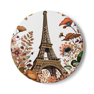 May include: A round coaster with a white background featuring a detailed illustration of the Eiffel Tower in brown and gold tones. The tower is surrounded by a wreath of colorful flowers and leaves in shades of orange, pink, and green.