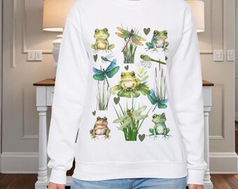 Könnte beinhalten: Weißer Pullover mit einem grünen Frosch- und Libellen-Design. Das Design zeigt mehrere Frösche und Libellen mit grünen Blättern und braunen Herzen.