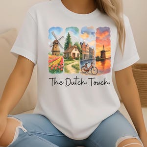 Może przedstawiać: Biała koszulka z akwarelowym wzorem przedstawiającym holenderskie wiatraki, domy, rower i pola tulipanów. Tekst "The Dutch Touch" jest wydrukowany poniżej. Koszulka jest wykonana z miękkiego materiału.