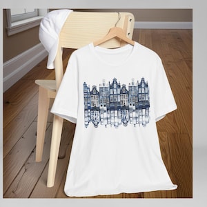Może przedstawiać: Biały t-shirt z krótkim rękawem z niebieskim nadrukiem graficznym przedstawiającym rząd kamienic nad kanałem z efektem odbicia lustrzanego. Gładka biała czapka z daszkiem jest przewieszona przez oparcie drewnianego krzesła.
