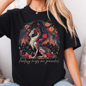 May include: Black t-shirt featuring a fantasy dragon design. The dragon is white with red and teal accents, surrounded by colorful flowers. The text "Fantasy keeps me grounded" is printed below the dragon.