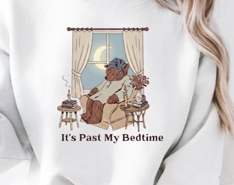 Può includere: Felpa bianca con un'illustrazione di un orso in pigiama seduto su una sedia vicino a una finestra con una luna crescente. Il testo "It's Past My Bedtime" è stampato sotto. Piccoli tavoli con libri e candele.