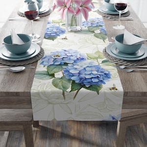 Pode incluir: Um caminho de mesa floral com um design em aquarela de hortênsias azuis e folhas verdes sobre um fundo creme. O caminho de mesa é decorado com abelhas e contornos de rosas brancas. A mesa está posta com tigelas, pratos e taças de vinho turquesa.