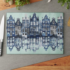 Könnte beinhalten: Eine blaue und weiße Aquarellillustration einer Reihe von Häusern, die sich in einem Kanal spiegeln. Die Häuser haben Giebeldächer und viele Fenster. Die Illustration ist auf einem hellblauen Hintergrund.