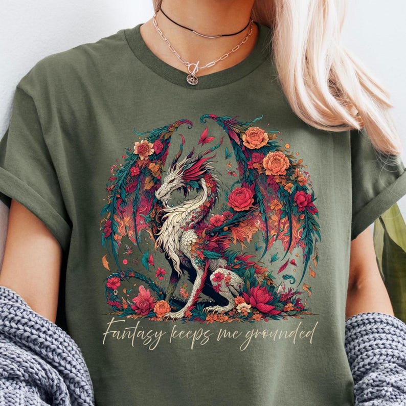 May include: Olive green t-shirt featuring a detailed illustration of a dragon with floral wings and accents. The dragon is white with red and pink details, surrounded by flowers. The text "Fantasy keeps me grounded" is below the dragon.