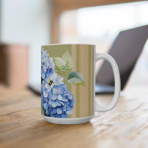 May include: White ceramic mug featuring a watercolor design of blue hydrangeas and a bee. The mug has a striped background in shades of green and tan. The handle is white and the mug is on a wooden surface.