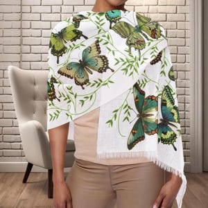 May include: White scarf with a butterfly print in shades of green, blue, and brown. The scarf has a fringed edge and is draped over the shoulders. The butterflies are interspersed with green leaf designs.
