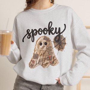 Può includere: Una felpa girocollo grigio chiaro con la scritta spooky in corsivo nero. Sotto il testo è presente una grafica di un fantasma realizzato con vari ritagli di tessuto scozzese beige e marrone con due bottoni neri al posto degli occhi.