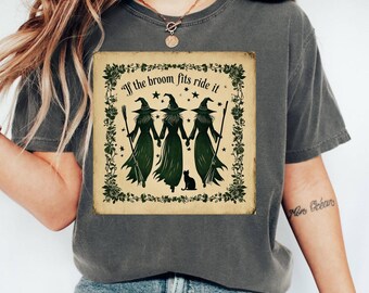 May include: Dark gray t-shirt featuring a vintage-style graphic. The graphic depicts three witches holding hands, with the text "If the broom fits ride it." A black cat is at the bottom of the image, and the design is framed by a floral border.