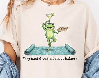 May include: Beige t-shirt featuring a cartoon frog balancing a margarita and a pizza box on a yoga mat. The frog is green with a white belly. The text "They said it was all about balance" is printed below the yoga mat.