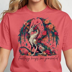 May include: Coral-colored t-shirt featuring a detailed illustration of a dragon with floral wings and accents. The dragon is white with red and green details, surrounded by flowers and butterflies. The text "Fantasy keeps me grounded" is below the dragon.